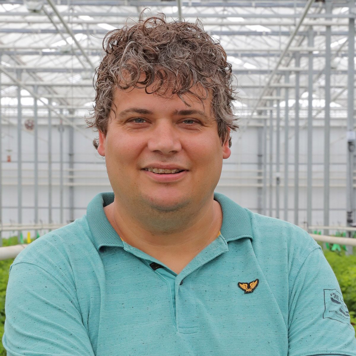 A person standing in a greenhouse with a structured metal framework. The person is wearing a light blue polo shirt with a small emblem on the left chest area. The greenhouse has a clear roof, and there are various plants visible in the background.
