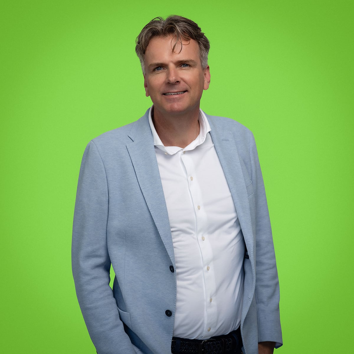 The image shows a person standing against a bright green background. The person is wearing a light blue blazer over a white shirt with a classic collar and buttons. The individual's face is not visible in the image.