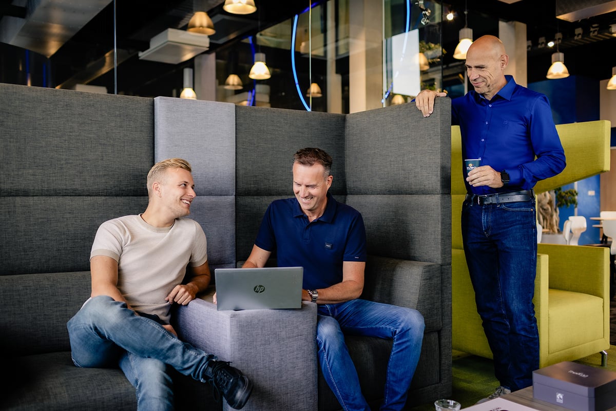 Three individuals are seated on a grey couch in an indoor setting with modern decor. Two are focused on a laptop, while the third, standing, is holding a beverage.