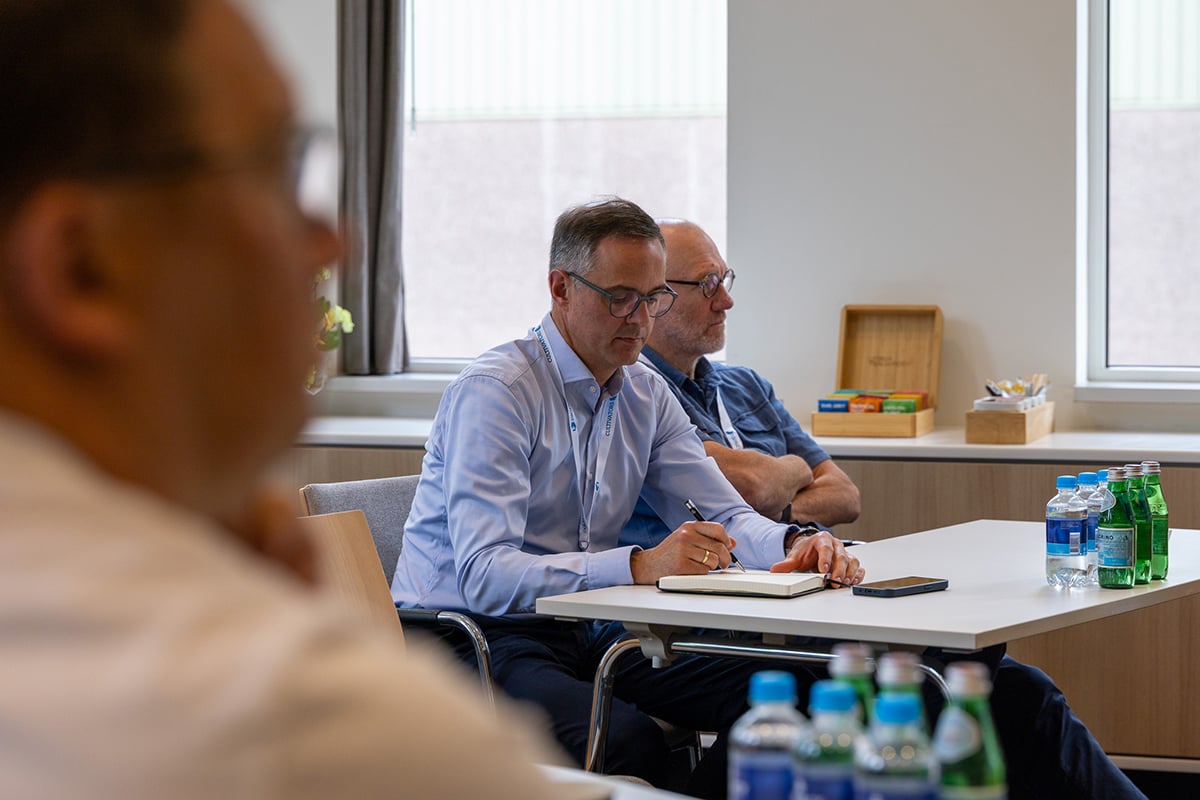 A professional setting where three individuals are seated around a table. The person in the foreground is wearing glasses and appears to be taking notes. Bottles of water are on the table, suggesting that the meeting could be lengthy.