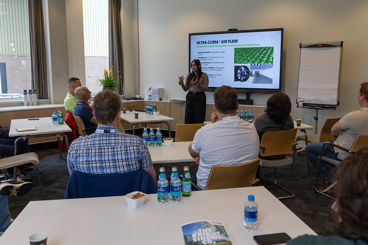 An indoor setting where a presentation is being given. A speaker stands in front of a class, presenting information on a screen that is displaying an image of a greenhouse. The audience, consisting of several individuals seated at tables, appears to be engaged with the presentation.