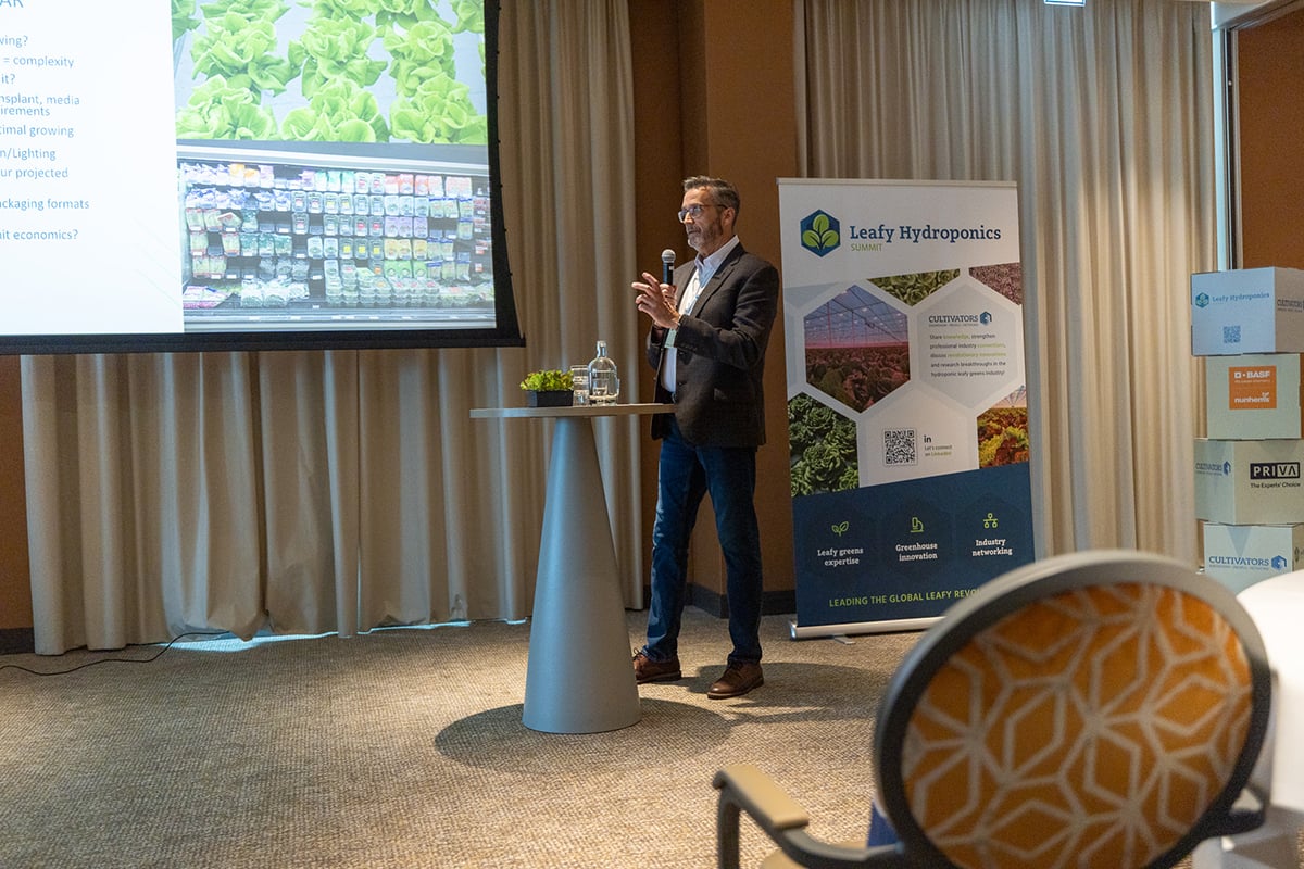 An indoor event where a presenter, whose face is blurred, is standing by a podium with a microphone, giving a presentation. Behind the presenter, there is a large screen displaying text and images related to hydroponics, specifically focused on leafy greens.
