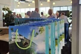 The image shows a group of people gathered in what appears to be a modern indoor space. They are standing behind a long, transparent barrier, which is adorned with a banner that reads 'GROWING WORLD'. The setting suggests an event or exhibition related to plant growth or gardening.