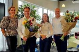 Four people are standing together in an indoor setting, each holding a bouquet of flowers. They are dressed in semi-formal attire, with the individuals in the center and to the right wearing white shirts, and the two on the left wearing patterned tops. The background suggests a casual event.