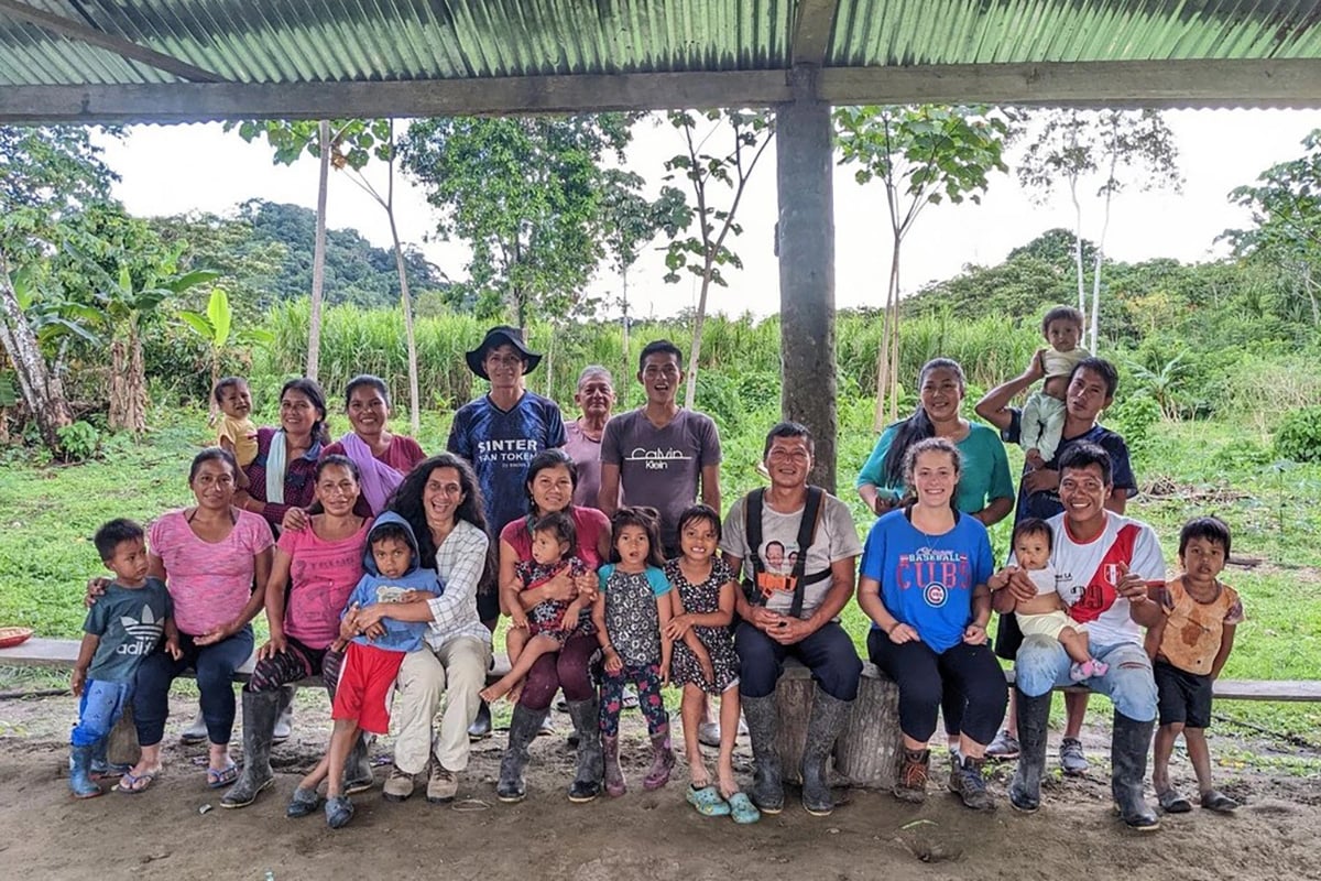 A group of people are gathered under a shelter, with some sitting and some standing, in a rural outdoor setting.