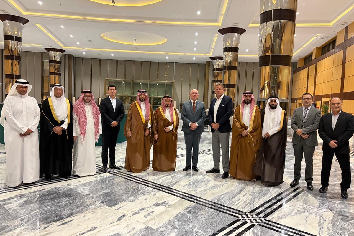 A group of individuals in a formal setting, possibly a business or diplomatic event. There are two rows of people, with the front row standing and the back row seated. The individuals are dressed in a mix of traditional and formal Western attire, suggesting a diverse gathering.