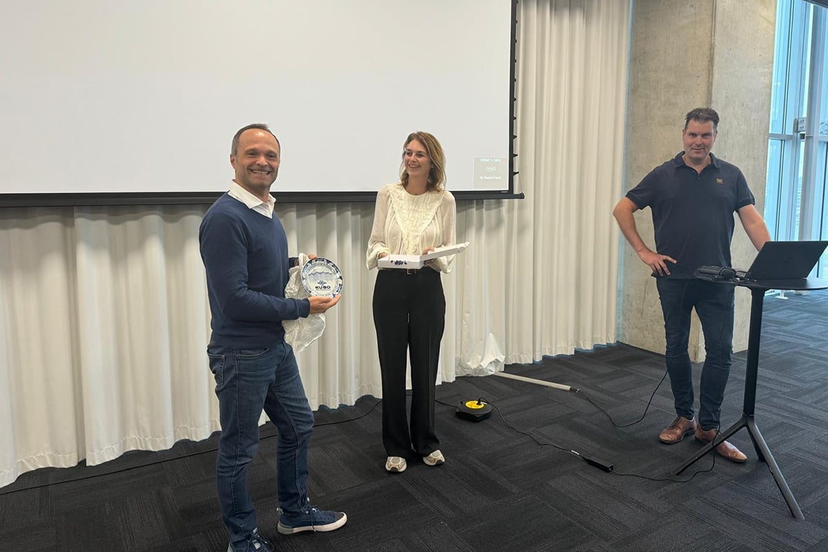 There are three individuals in a room with a white screen and a white curtain in the background. The person on the left is wearing a blue sweater and jeans, holding a white object. The person in the middle is also holding a white object.