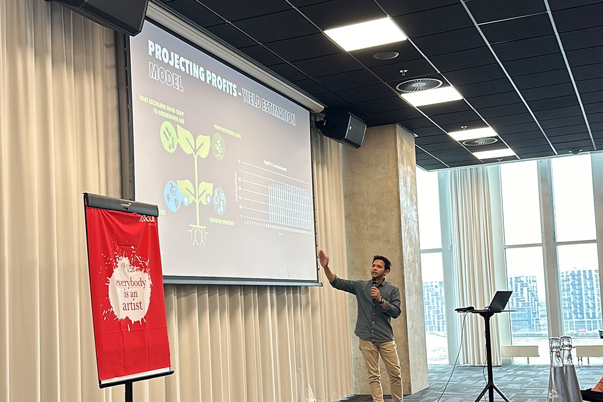 A man giving a presentation in a conference room. He is standing in front of a projection screen displaying charts and graphs. There is a speaker at his right, and a red banner with the text 'everyone is an artist' is visible on the left.