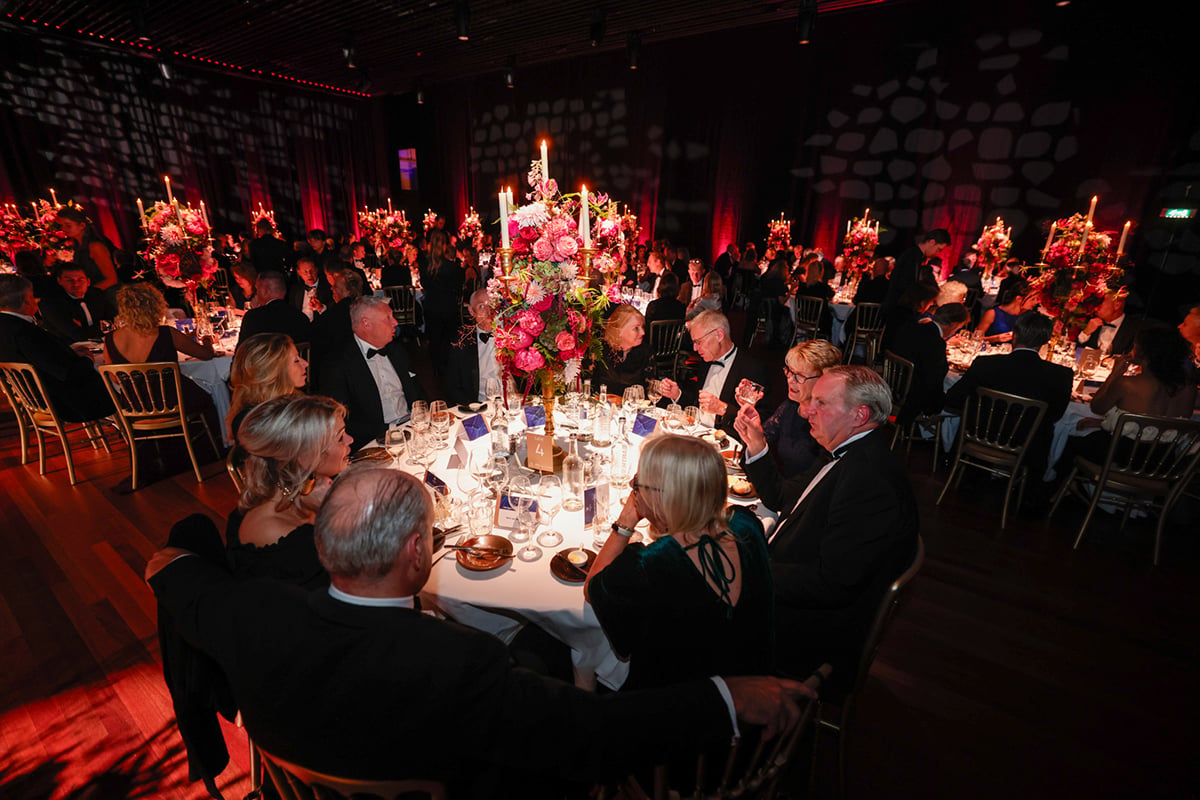 A formal dinner event inside a dimly lit venue with a chandelier. Attendees are seated around a long table, adorned with elegant table settings and floral centerpieces. The guests are dressed in formal attire, and the atmosphere appears to be intimate and upscale.