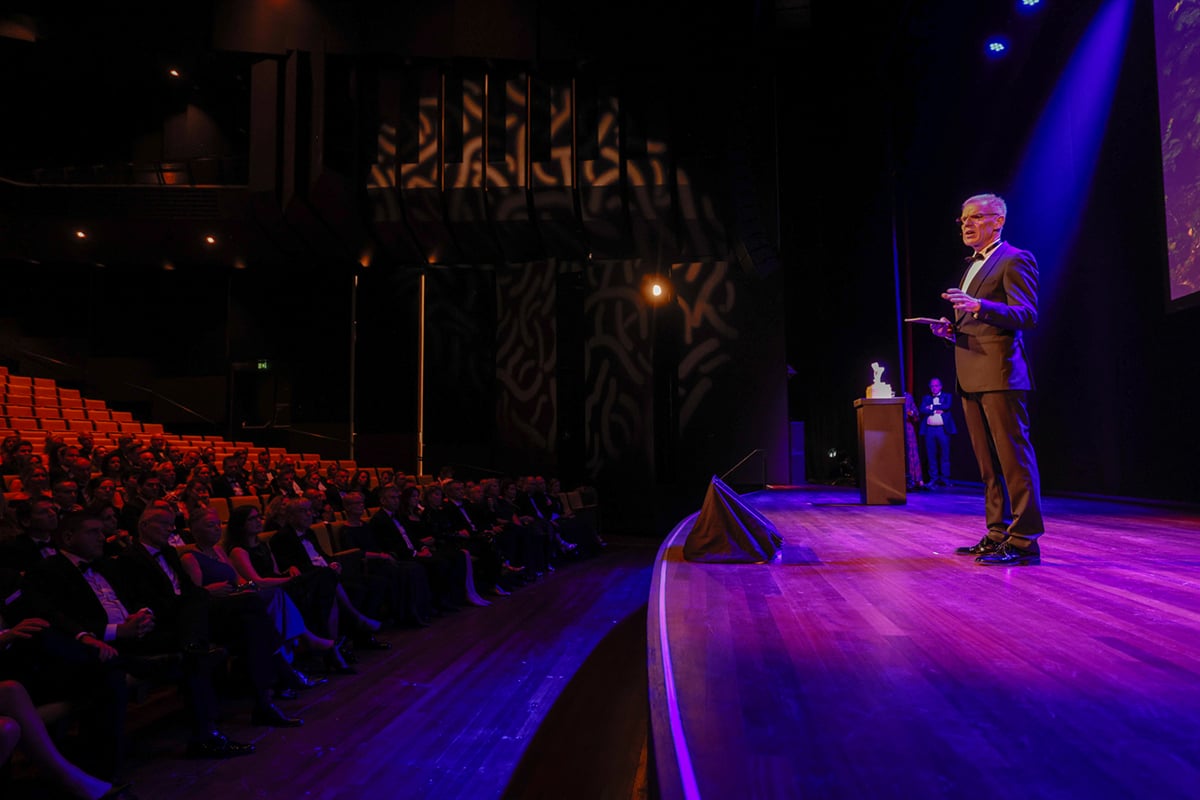 A man standing at a podium with a microphone, addressing an audience in a dimly lit indoor setting. The audience is seated in rows, facing the speaker, and appears attentive. The stage has a curved edge, and the setting has a serious, formal atmosphere.