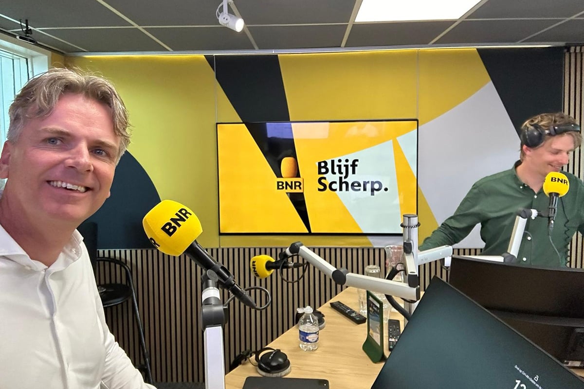An indoor setting, likely a radio studio with two individuals. One person is seated at a desk with a laptop, a microphone, and other equipment, while the other stands behind the desk with a microphone. The room has a modern design with yellow and black geometric patterns.