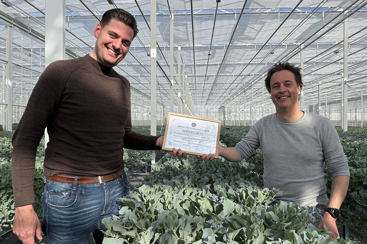 Two people standing among green plants in a greenhouse, one holding a certificate.