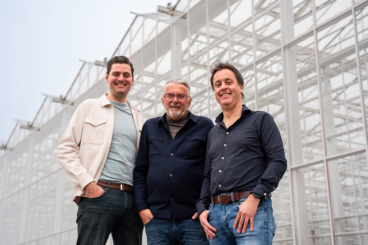 Three people standing side by side at a construction site, with a large unfinished metal framework structure in the background under an overcast sky.