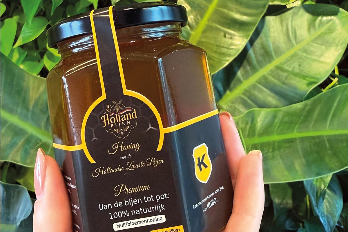 The image shows a hand holding a jar of Hollanda Hening honey. The jar is designed to look like a traditional honey pot and has a label with the product name, a description in multiple languages, and a logo. The background features a green leafy plant.