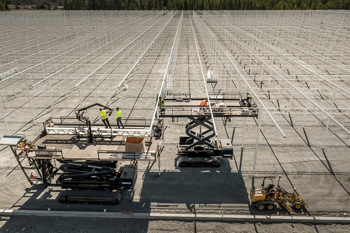 A large outdoor area, likely a solar farm, with rows of solar panels installed on the ground. There are several pieces of industrial equipment, including a crane, a concrete mixer, and what appears to be a worker in high-visibility clothing.