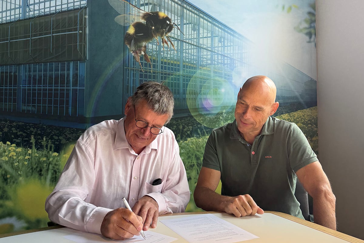 The image shows two men sitting at a table, each writing on a piece of paper. The man on the left is wearing a white shirt and glasses, while the man on the right is wearing a green polo shirt. The background features a large greenhouse, and there's a bee on the left side of the image.