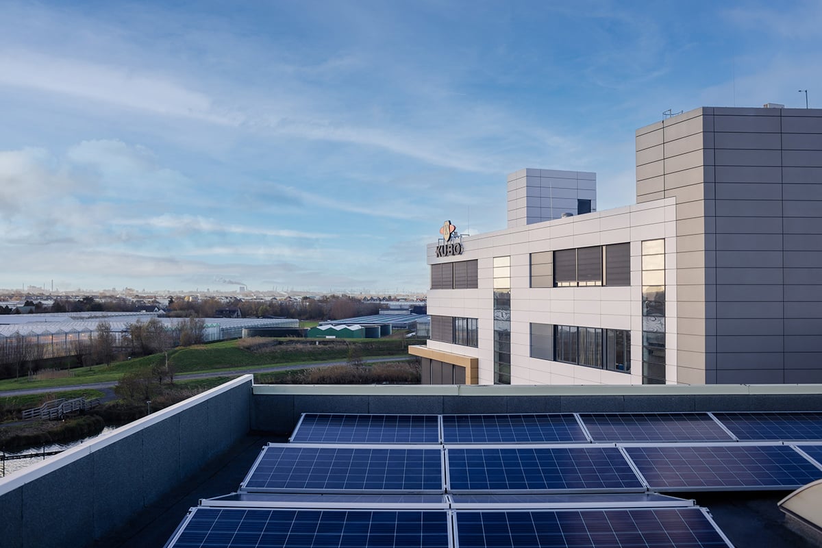 The image shows a modern building with a flat roof covered in solar panels. The building is composed of a series of rectangular structures with large windows. In the background, there is a body of water and some bare trees. The sky is partly cloudy.
