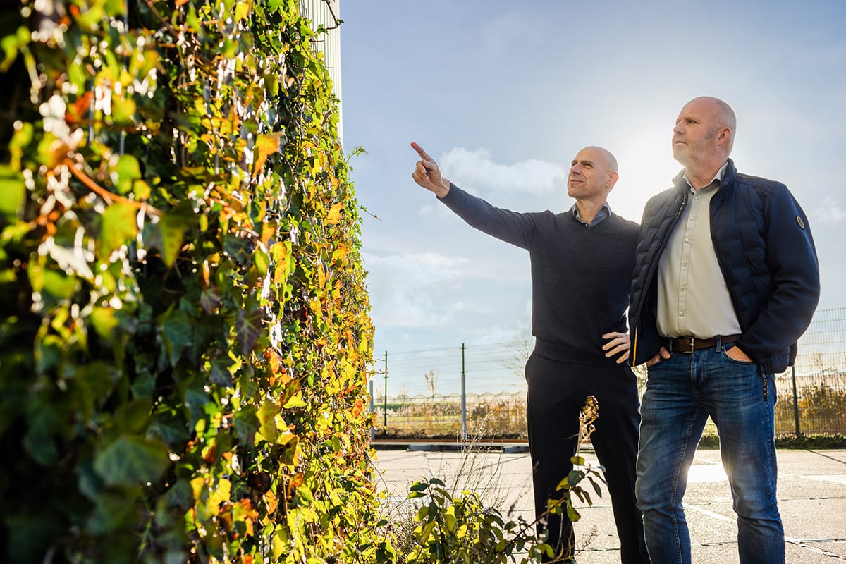 Two individuals are standing near a living wall covered with greenery. One person is pointing towards the sky, while the other stands beside them, observing.
