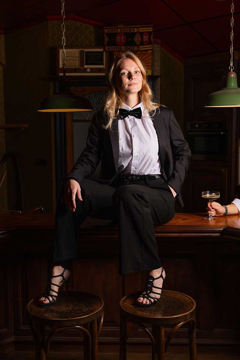 A woman dressed in formal wear is seated on a stool at a counter with a glass of wine in hand, in a room with vintage decor.