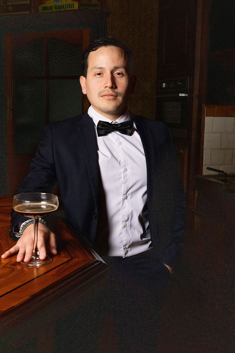 Aman standing behind a bar counter, dressed in a formal black suit with a white shirt and a black bow tie. He is holding a martini glass with a clear liquid, presumably a martini, in his hand. The background features a dimly lit bar setting with a closed glass door.