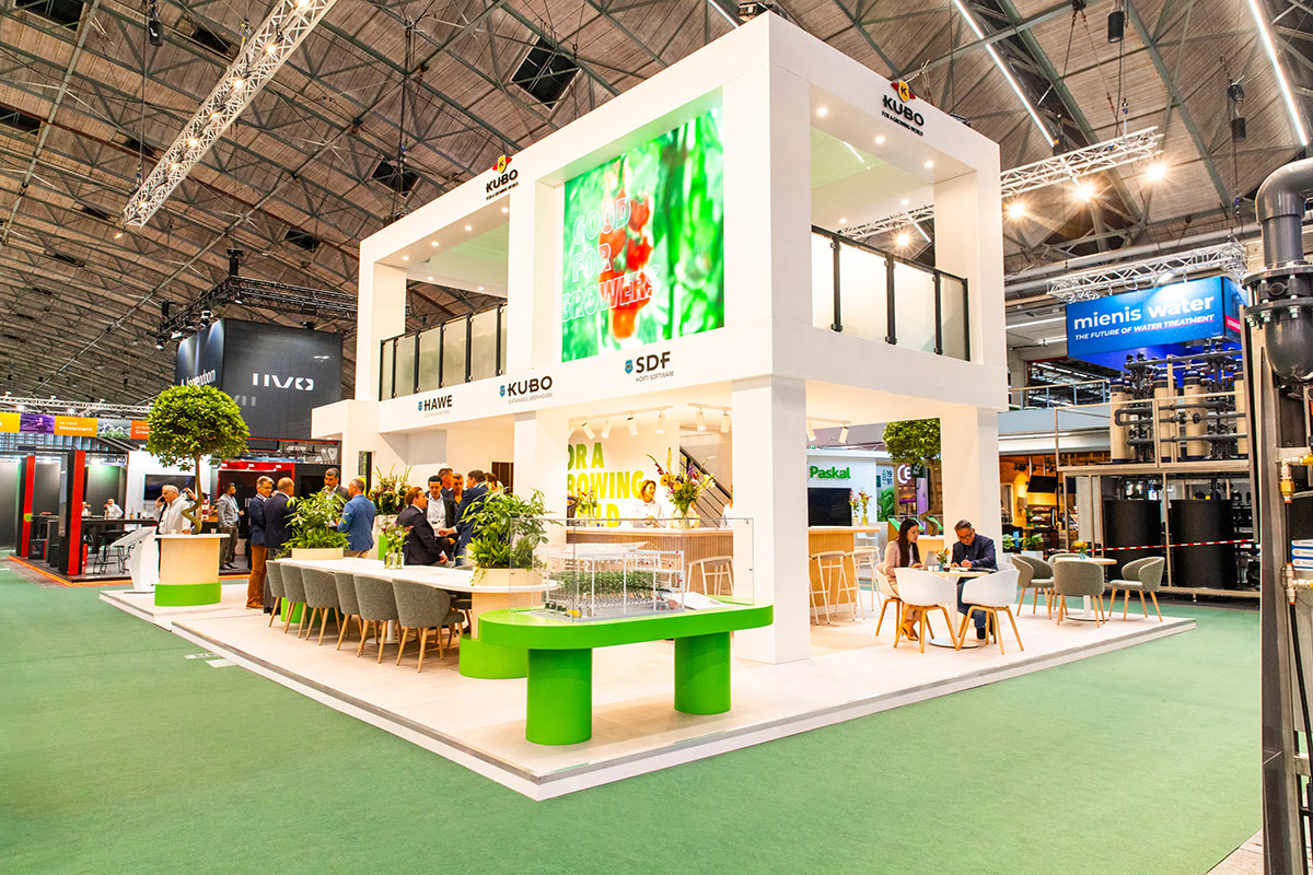 Een moderne expositieruimte met een witte stand met een groen en wit kleurenschema. Er zitten en staan mensen bij de stand, die een groot scherm heeft met kleurrijke afbeeldingen. De stand wordt omringd door andere beursstands en heeft een groene vloerbedekking.