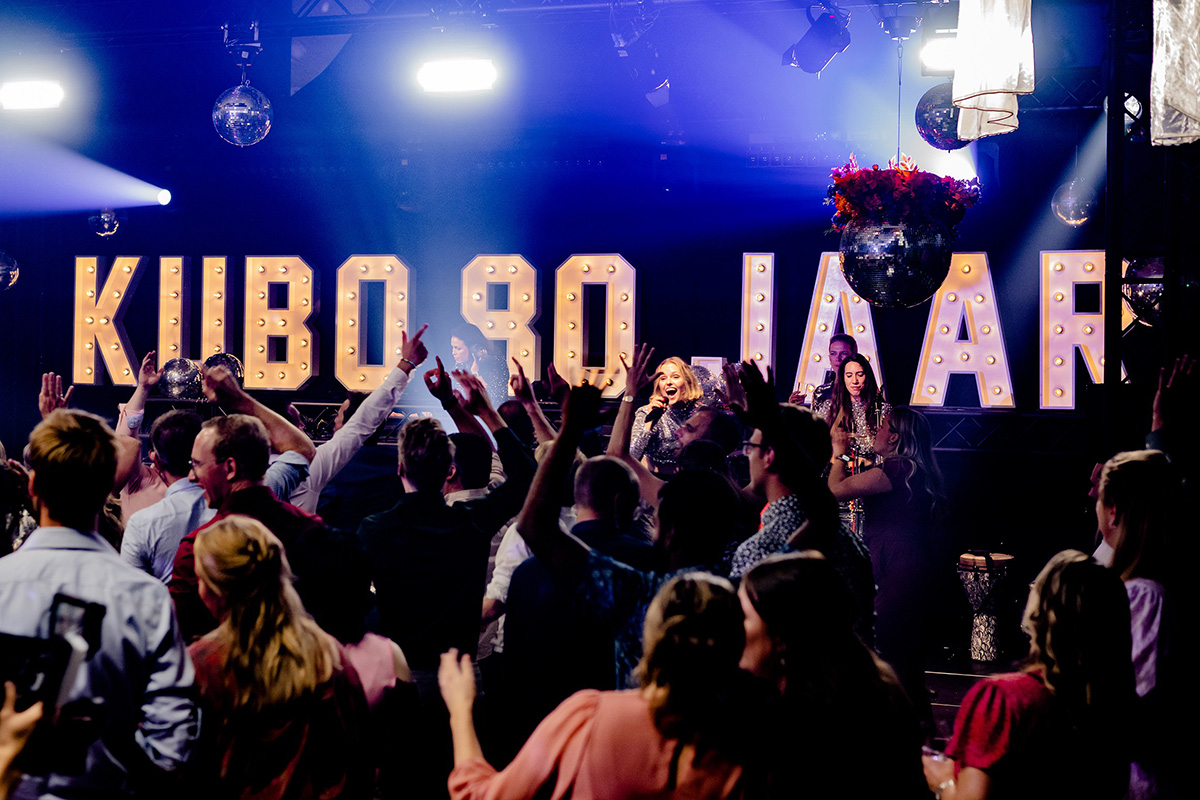 Group of people celebrating at an event or nightlife venue, with colorful lighting, raised hands, and a large illuminated letter display in the background.