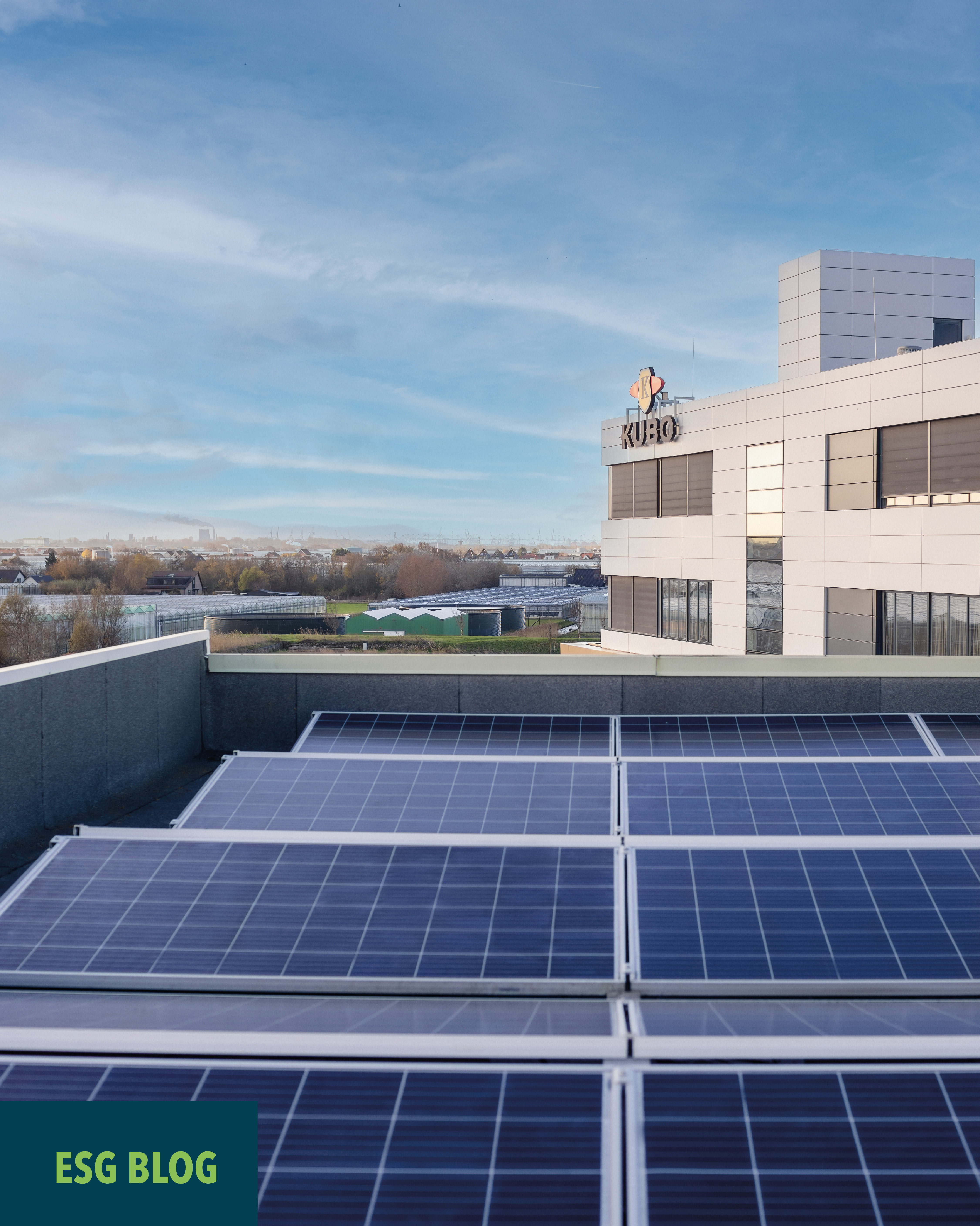 Solar panels installed on a flat rooftop of a modern building, overlooking an open landscape under a blue sky with light clouds.